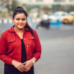 Outdoor close up Portrait of young brunette Asian/ Indian girl wearing red denim shirt and smiling while looking at camera in city.